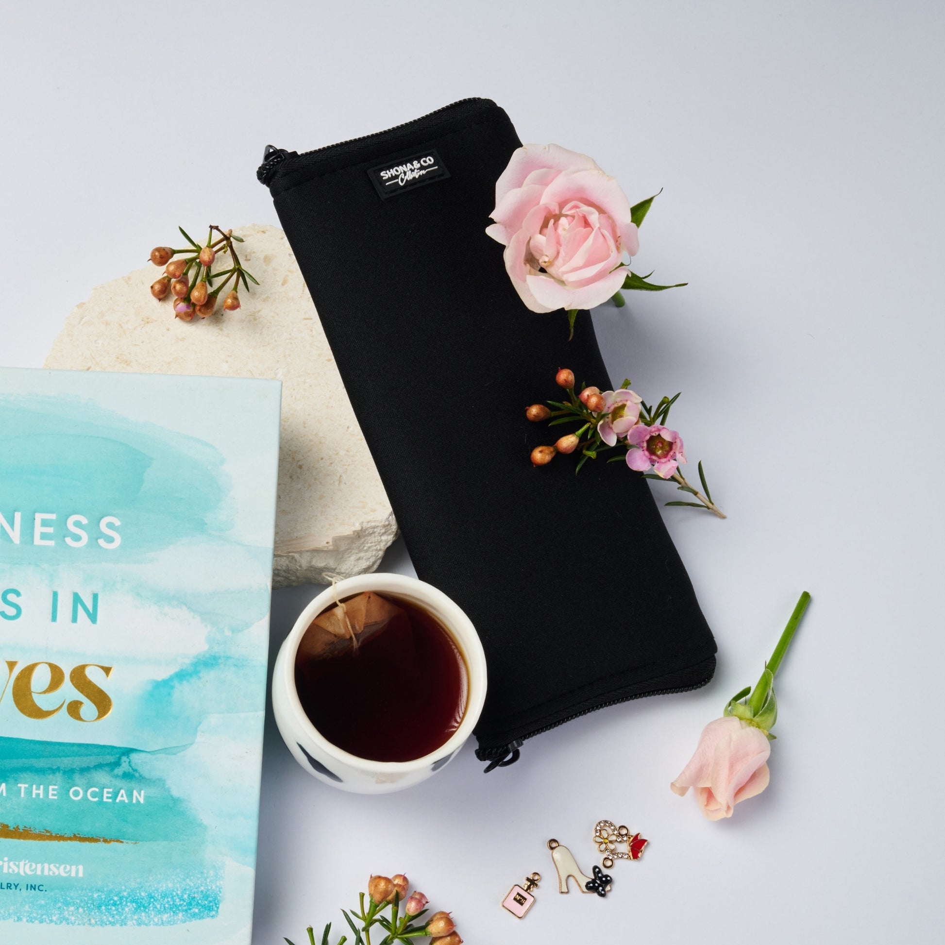 Black pouch, cup of tea, book, and flowers on a light gray background
